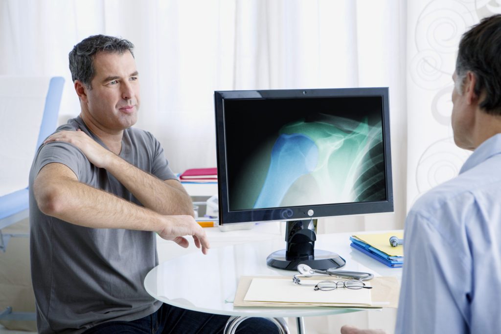 Man holding his shoulder in pain, sitting behind an xray and a doctor